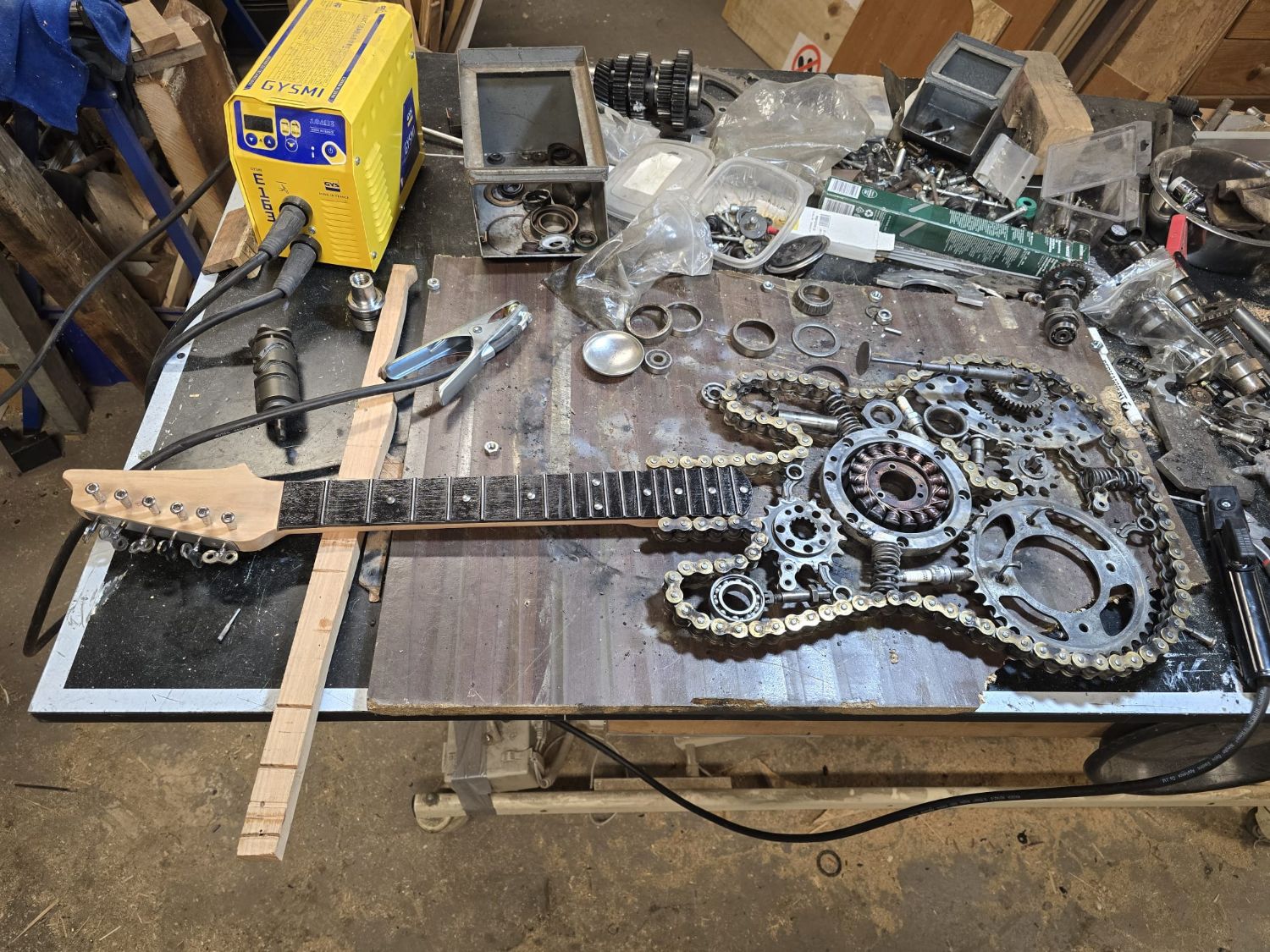 La guitare en cours d'assemblage sur l'établi, entourée de chaînes, pignons et engrenages de moto